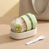 A small portable yogurt container with a light green lid and base, shown partially filled with yogurt, oats, and kiwi slices, resting on a wooden surface. A white spoon is placed in front, and a soft beige tote bag is blurred in the background.