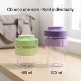 Two clear reusable travel cups on a light wood surface, one with a light green lid and band labeled 480 ml and the other with a lavender purple lid and band labeled 570 ml. Text at the top reads 'Choose one size - Sold individually'.