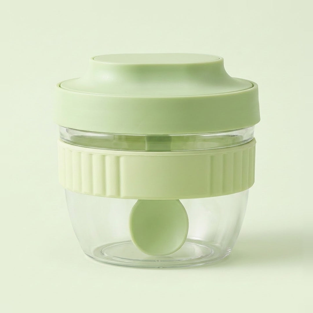 A small clear plastic food chopper jar with a light green lid, a matching green inner blade, and a ribbed light green collar, shown on a pale green background.