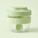 A small clear plastic food chopper jar with a light green lid, a matching green inner blade, and a ribbed light green collar, shown on a pale green background.