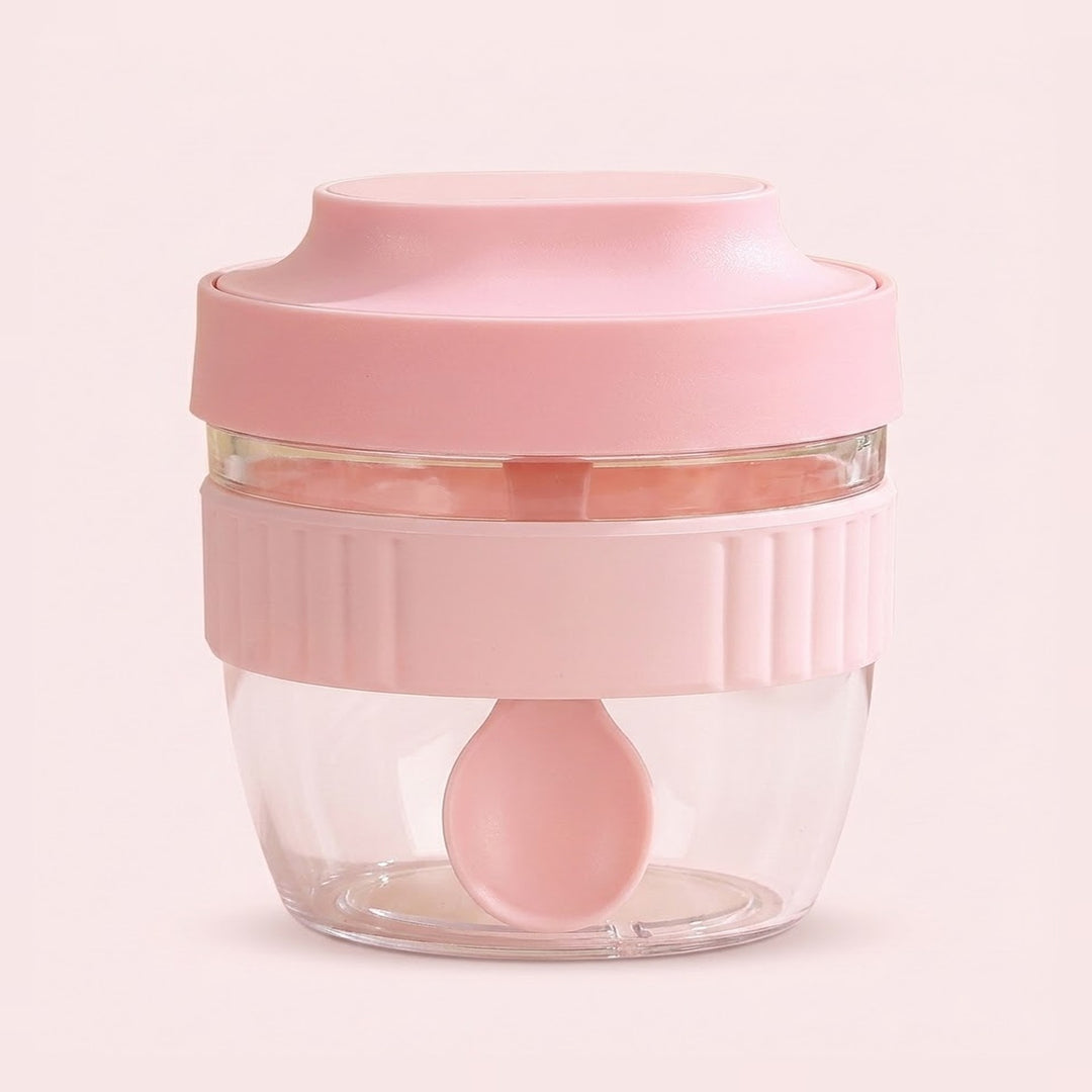 A small pink food chopper container with a clear plastic bowl, a pink lid, a ribbed pink handle, and a matching pink spoon inside, set against a solid pale pink background.