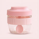 A small pink food chopper container with a clear plastic bowl, a pink lid, a ribbed pink handle, and a matching pink spoon inside, set against a solid pale pink background.