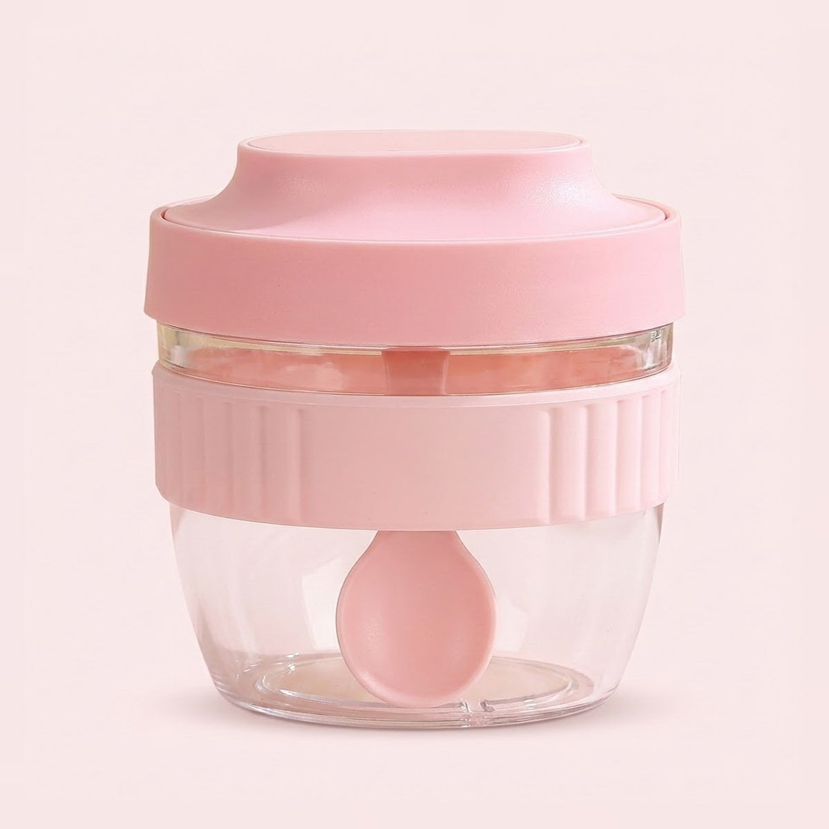 A small pink food chopper container with a clear plastic bowl, a pink lid, a ribbed pink handle, and a matching pink spoon inside, set against a solid pale pink background.