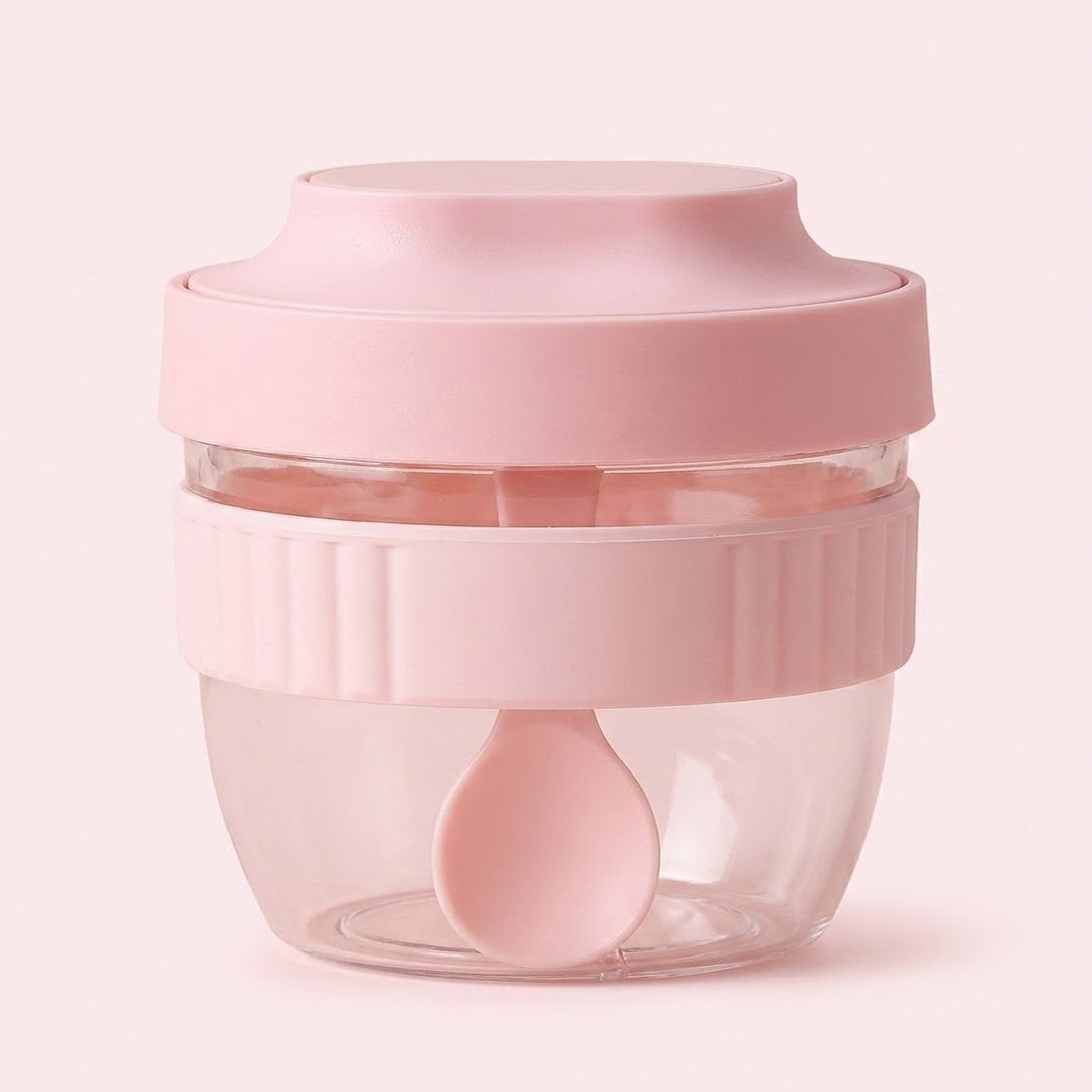 A small pink food storage container with a clear plastic bowl, a ribbed pink lid, and a matching pink spoon inside, set against a solid pale pink background.