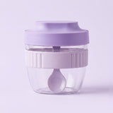 A small portable blender cup with a lavender purple lid, a white ribbed middle band, and a clear plastic base containing a matching lavender spoon, set against a solid light purple background.