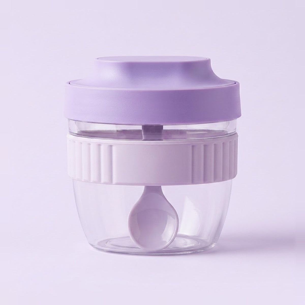 A small portable blender cup with a lavender purple lid, a white ribbed middle band, and a clear plastic base containing a matching lavender spoon, set against a solid light purple background.