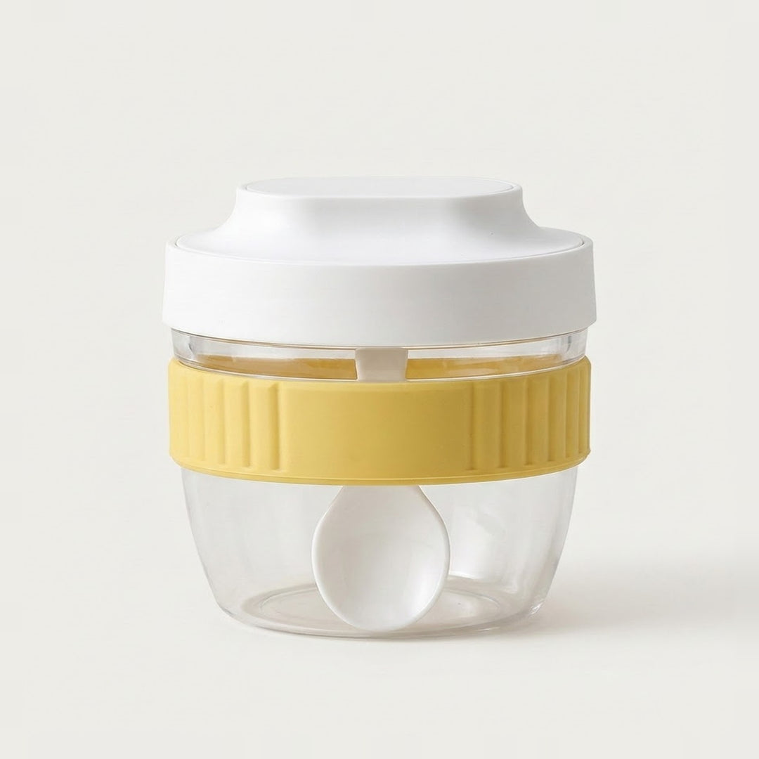 A clear plastic food storage container with a white lid, a yellow ribbed silicone band, and a white spoon inside, set against a plain off-white background.