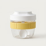 A clear plastic food storage container with a white lid, a yellow ribbed silicone band, and a white spoon inside, set against a plain off-white background.