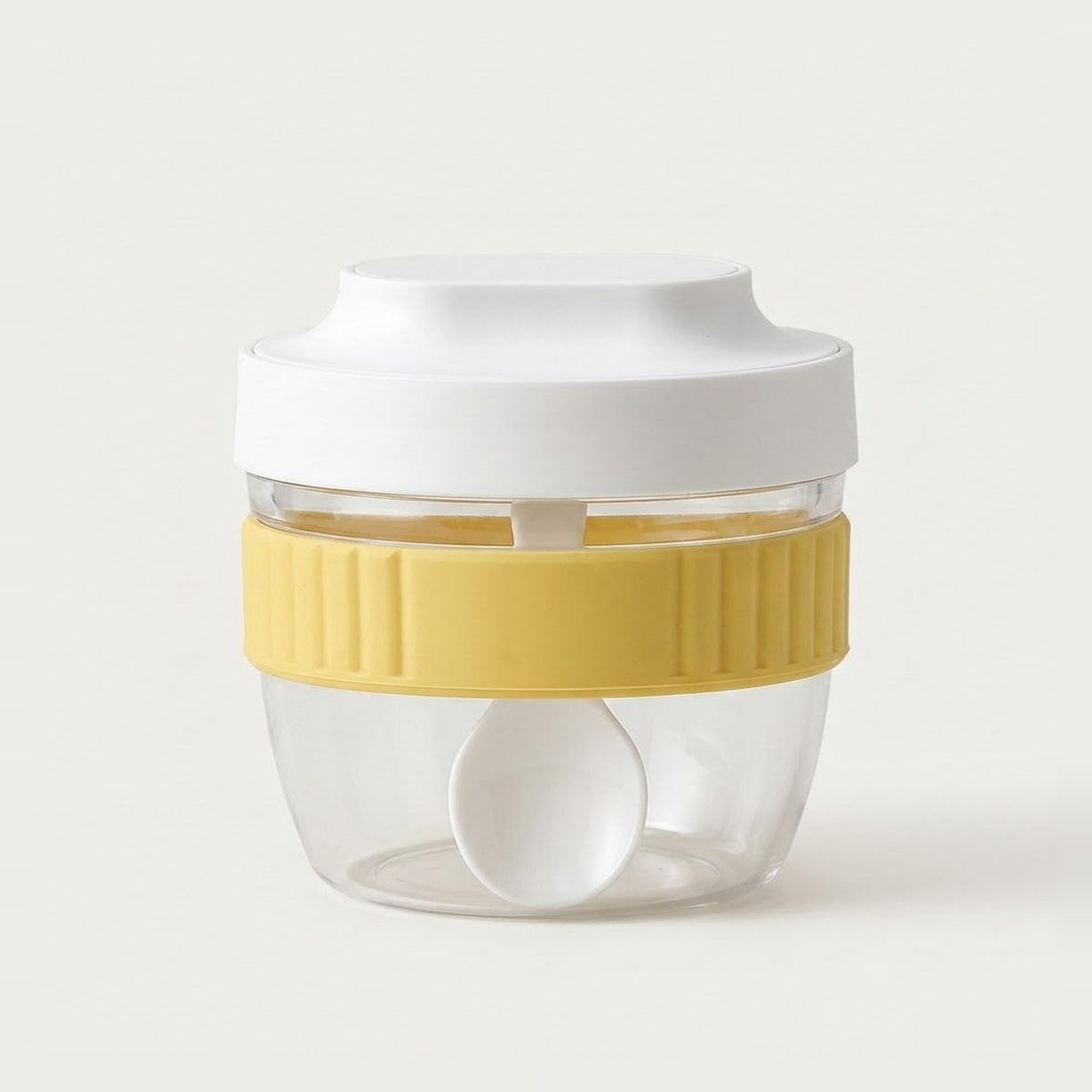 A clear plastic food storage container with a white lid, a yellow ribbed silicone band, and a white spoon inside, set against a plain off-white background.