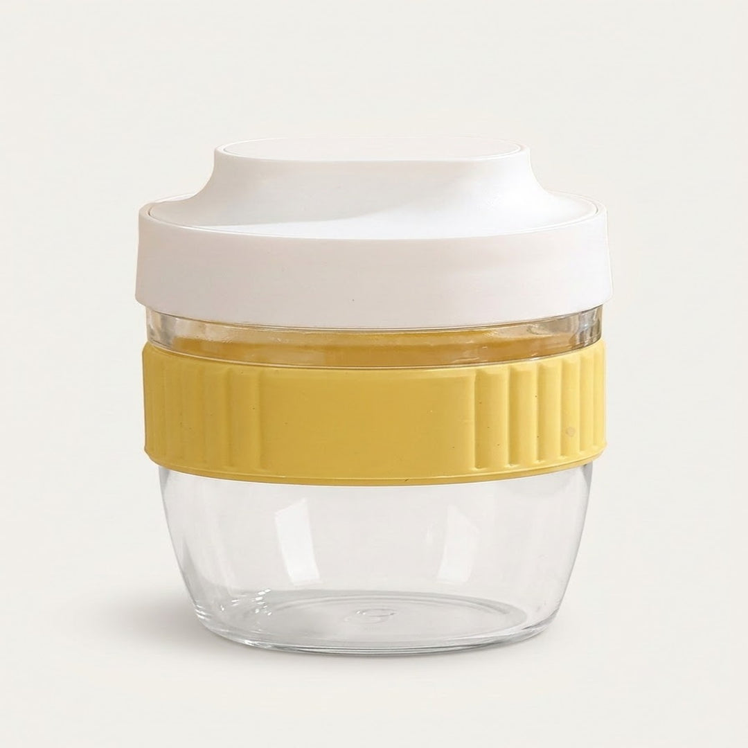 A reusable glass food container with a clear body, a yellow ribbed silicone grip band, and a white lid, shown on a light off-white background.