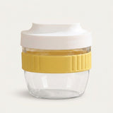 A reusable glass food container with a clear body, a yellow ribbed silicone grip band, and a white lid, shown on a light off-white background.