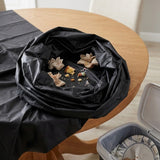 A black table-top trash bag draped over a round wooden table, with crumpled paper and food debris visible inside. An open gray trash can with a liner sits in the lower right corner.