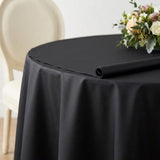 A black round tablecloth draped over a round table, with a matching rolled section on top and a floral centerpiece in the corner. A cream-colored upholstered chair is visible in the background against a light neutral wall.