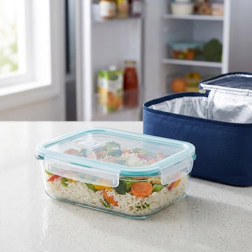 A clear glass food storage container with a light blue lid, filled with rice and vegetables, sits on a kitchen counter next to a navy blue insulated lunch bag with a silver lining.