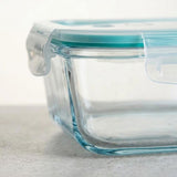 A close-up of a clear glass food storage container with a translucent aqua lid and a clear plastic latch, resting on a light gray surface.