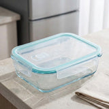 A clear glass storage container with a light blue silicone seal and lid, shown on a kitchen countertop with a folded cloth and a blurred refrigerator in the background.