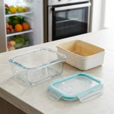 A set of glass food storage containers on a kitchen countertop, including a clear glass container with a light blue silicone lid, a beige container, and a clear glass container with a clear lid.