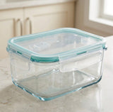 Clear glass storage container with a translucent aqua lid and handle, shown on a kitchen countertop with cabinets and a window in the background.