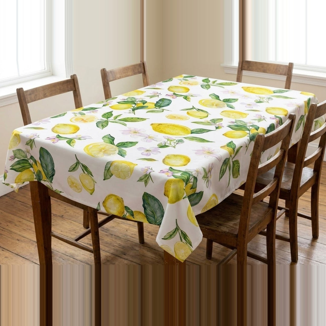 A rectangular table covered with a white tablecloth printed with a lemon and leaf pattern, surrounded by wooden dining chairs in a bright room with windows and wood flooring.