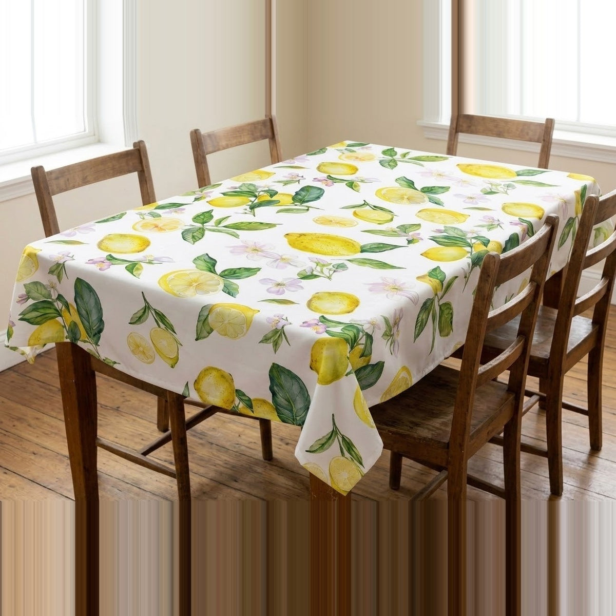 A rectangular table covered with a white tablecloth printed with a lemon and leaf pattern, surrounded by wooden dining chairs in a bright room with windows and wood flooring.