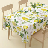 A rectangular table covered with a white tablecloth printed with yellow lemons, green leaves, and small pale pink flowers, shown with light wood chairs in a clean studio setting.