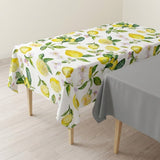 A rectangular table covered with a white tablecloth featuring a yellow lemon and green leaf pattern, shown in a room with light wood flooring and a second gray tablecloth partially visible on the right.