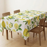 A rectangular table covered with a white tablecloth printed with a bright yellow lemon and green leaf pattern, shown in a dining room setting with wooden chairs and a light wood floor.