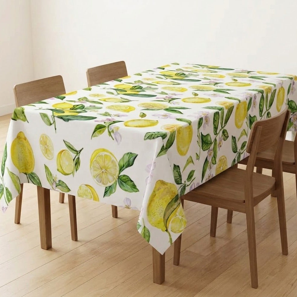 A rectangular table covered with a white tablecloth printed with a bright yellow lemon and green leaf pattern, shown in a dining room setting with wooden chairs and a light wood floor.