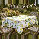 A rectangular outdoor dining table covered with a white tablecloth printed with yellow lemons and green leaves, surrounded by wooden chairs in a garden setting with bunting in the background.