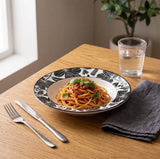A round ceramic plate with a black floral pattern on a white base, filled with spaghetti and garnished with basil, set on a wooden table with a fork, knife, folded napkin, and a glass of water.