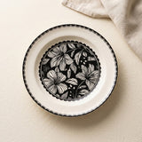 A round white ceramic plate with a black scalloped rim and a black-and-white floral pattern in the center, photographed from above on a light beige textured surface with a folded cream cloth in the corner.