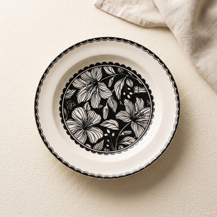 A round white ceramic plate with a black scalloped rim and a black-and-white floral pattern in the center, photographed from above on a light beige textured surface with a folded cream cloth in the corner.