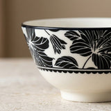 A close-up of a white ceramic bowl featuring a bold black floral pattern and a scalloped black band near the base, set on a light textured surface with a soft beige background.