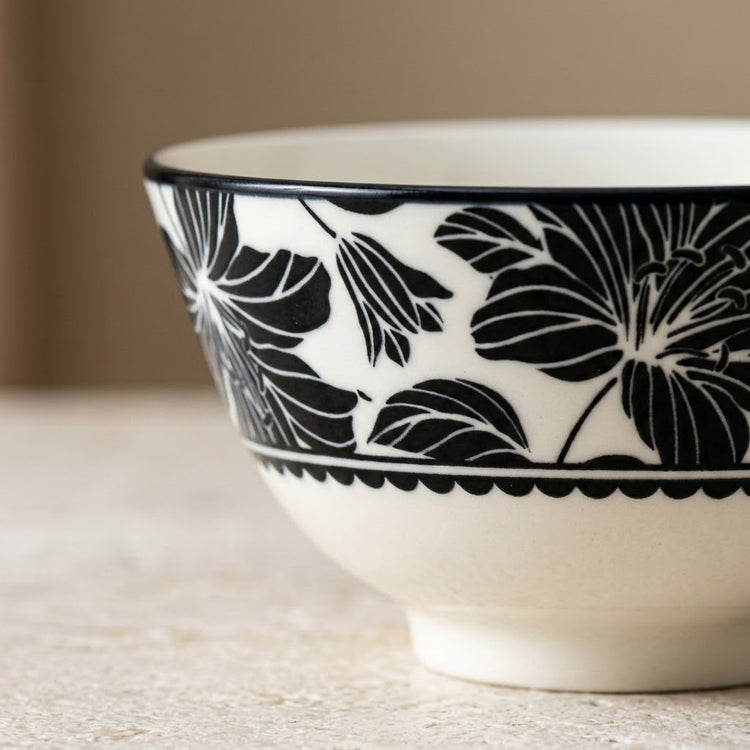 A close-up of a white ceramic bowl featuring a bold black floral pattern and a scalloped black band near the base, set on a light textured surface with a soft beige background.