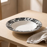 A wide white ceramic bowl with a black floral border pattern, resting on a light wood table next to a beige linen napkin.
