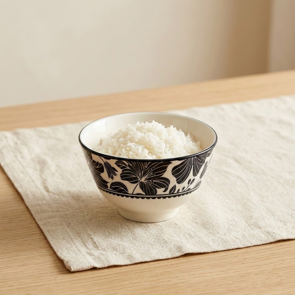 A ceramic bowl filled with white rice, decorated with a black floral pattern, resting on a beige linen placemat on a light wood table.