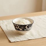 A ceramic bowl filled with white rice, decorated with a black floral pattern, resting on a beige linen placemat on a light wood table.