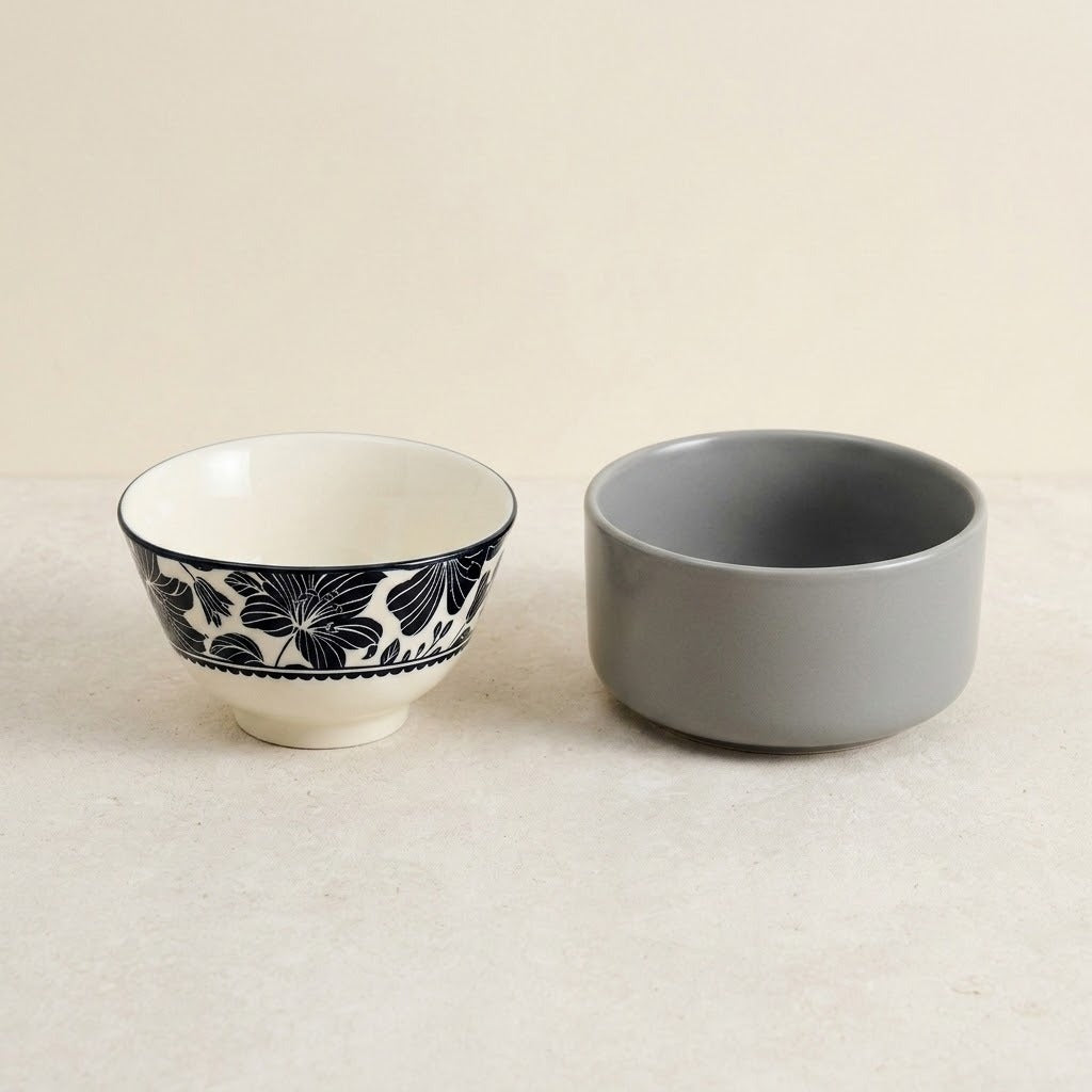 Two ceramic bowls on a light beige surface against a matching neutral background. One bowl is white with a black floral pattern around the rim, and the other is a solid matte gray.