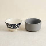 Two ceramic bowls on a light beige surface against a matching neutral background. One bowl is white with a black floral pattern around the rim, and the other is a solid matte gray.