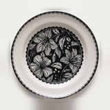 A round white ceramic plate with a black scalloped rim and a central black floral pattern, photographed from directly above on a white background.