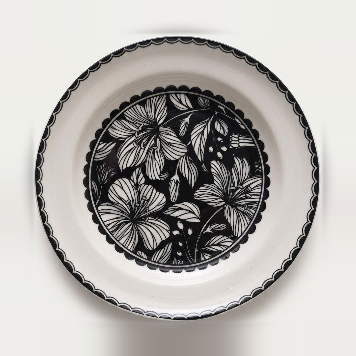A round white ceramic plate with a black scalloped rim and a central black floral pattern, photographed from directly above on a white background.