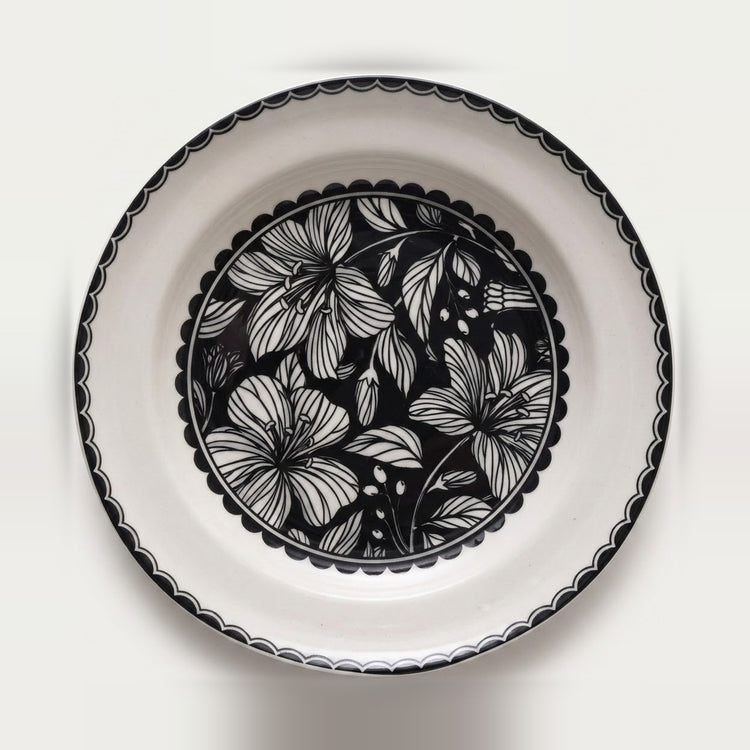 A round white ceramic plate with a black scalloped rim and a central black floral pattern, photographed from directly above on a white background.
