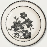 An off-white ceramic plate featuring a black floral design in the center and a black scalloped rim, shown against a plain light background.
