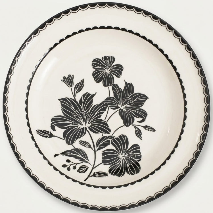 An off-white ceramic plate featuring a black floral design in the center and a black scalloped rim, shown against a plain light background.