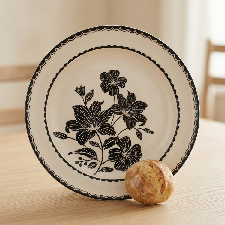 A round ceramic plate with a black floral and scalloped border design, shown on a light wooden table with a small bread roll in front of it.