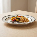 A round white ceramic plate with a black floral pattern and scalloped rim, shown on a light wood table with food on it.