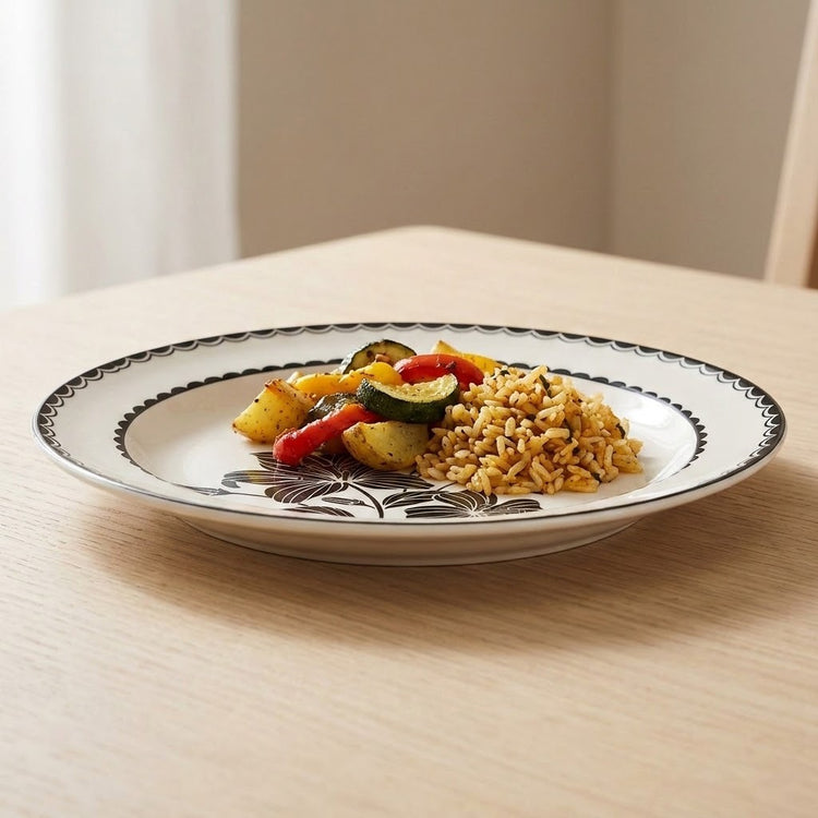 A round white ceramic plate with a black floral pattern and scalloped rim, shown on a light wood table with food on it.