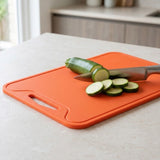 An orange rectangular plastic cutting board with a handle, shown on a light countertop with a zucchini being sliced with a knife. The background shows a softly blurred kitchen setting.
