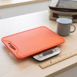An orange rectangular cutting board with a handle, shown on a digital kitchen scale on a light wood table, with a gray mug and a wooden ruler nearby.