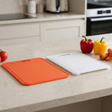 Two plastic cutting boards, one orange and one white, displayed on a light beige kitchen countertop with fresh vegetables nearby.